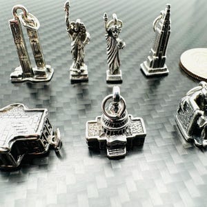 May include: A collection of silver-toned miniature charms depicting iconic American landmarks, including the Twin Towers, the Statue of Liberty, the Empire State Building, the Capitol Building, and the Lincoln Memorial. The charms are displayed on a textured surface.