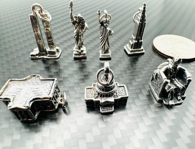 May include: A collection of silver-toned miniature charms depicting iconic American landmarks, including the Twin Towers, the Statue of Liberty, the Empire State Building, the Capitol Building, and the Lincoln Memorial. The charms are displayed on a textured surface.