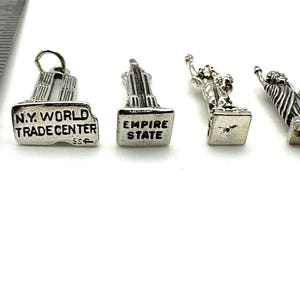 May include: Four silver-toned miniature charms. The charms depict the Twin Towers with "N.Y. WORLD TRADE CENTER" text, the Empire State Building with "EMPIRE STATE" text, and two Statue of Liberty figures. The charms are on a white surface.