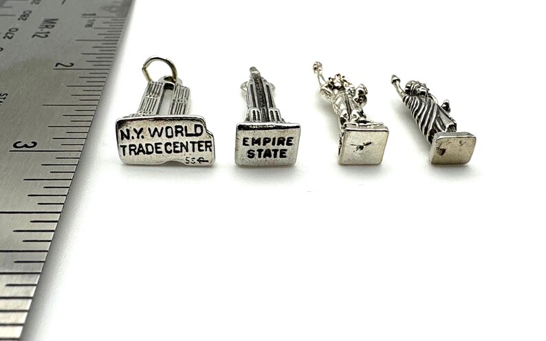 May include: Four silver-toned miniature charms. The charms depict the Twin Towers with "N.Y. WORLD TRADE CENTER" text, the Empire State Building with "EMPIRE STATE" text, and two Statue of Liberty figures. The charms are on a white surface.