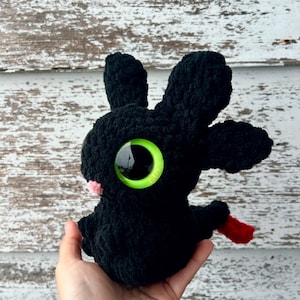 K&ouml;nnte beinhalten: Handgefertigtes schwarzes H&auml;keldrachen-Pl&uuml;schtier mit gro&szlig;en gr&uuml;nen Augen und einer rosa Nase. Der Drache hat eine rote Zunge und wird in einer Hand gehalten. Das Pl&uuml;schtier hat gro&szlig;e Ohren und ein weiches, strukturiertes Aussehen.