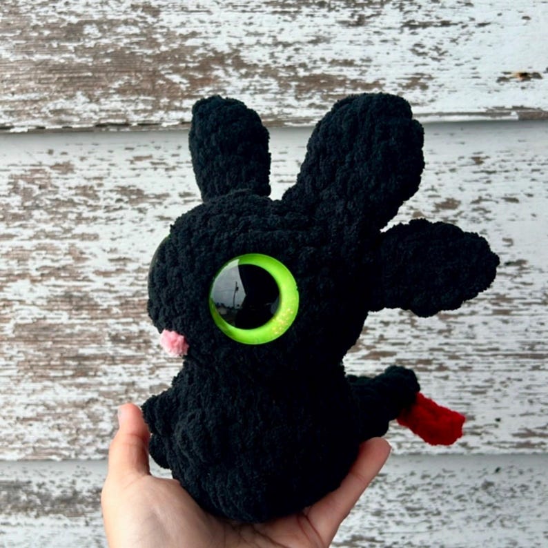 K&ouml;nnte beinhalten: Handgefertigtes schwarzes H&auml;keldrachen-Pl&uuml;schtier mit gro&szlig;en gr&uuml;nen Augen und einer rosa Nase. Der Drache hat eine rote Zunge und wird in einer Hand gehalten. Das Pl&uuml;schtier hat gro&szlig;e Ohren und ein weiches, strukturiertes Aussehen.