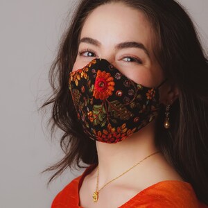 Op de afbeelding: Een vrouw draagt ​​een zwart gezichtsmasker met kleurrijke bloemenborduurwerk. Het masker heeft een gouden accent en de vrouw draagt ​​een gouden ketting met een klein hanger. Ze draagt ​​een feloranje top.