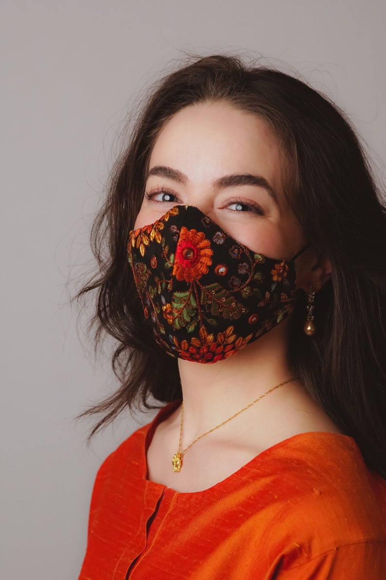 Op de afbeelding: Een vrouw draagt ​​een zwart gezichtsmasker met kleurrijke bloemenborduurwerk. Het masker heeft een gouden accent en de vrouw draagt ​​een gouden ketting met een klein hanger. Ze draagt ​​een feloranje top.