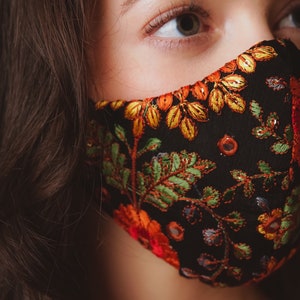 Op de afbeelding: Een zwart gezichtsmasker met kleurrijke bloemenborduurwerk. Het masker heeft een donkere achtergrond met oranje, gele, groene en rode bloemenpatronen.