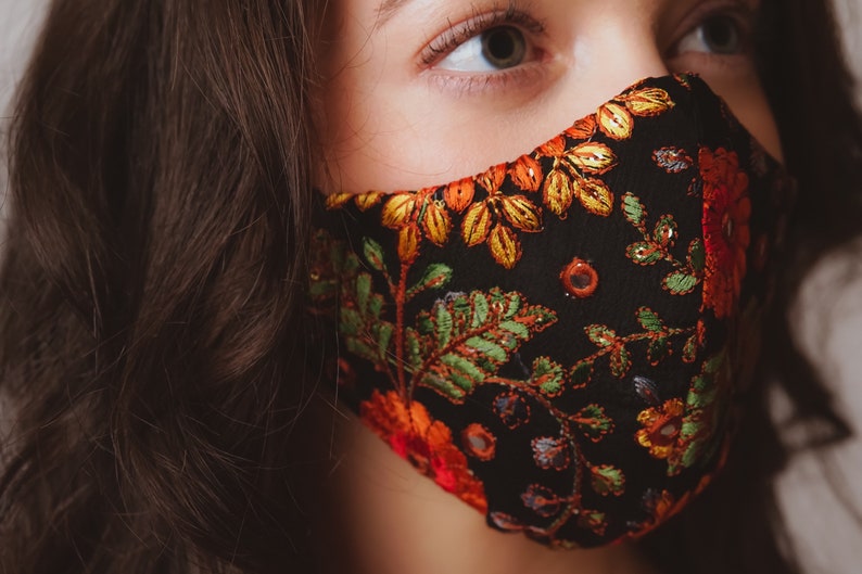 Op de afbeelding: Een zwart gezichtsmasker met kleurrijke bloemenborduurwerk. Het masker heeft een donkere achtergrond met oranje, gele, groene en rode bloemenpatronen.