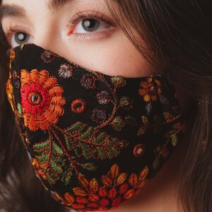 Op de afbeelding: Een zwart gezichtsmasker met een kleurrijk borduurwerk van bloemen. Het borduurwerk bevat rode, oranje, gele, groene en blauwe bloemen en bladeren.