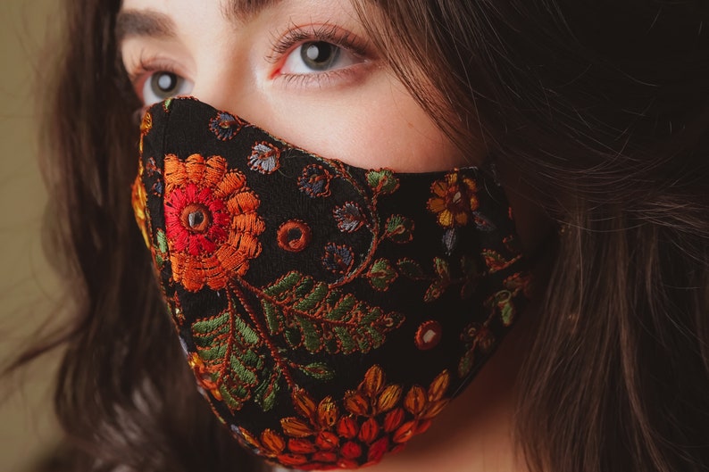 Op de afbeelding: Een zwart gezichtsmasker met een kleurrijk borduurwerk van bloemen. Het borduurwerk bevat rode, oranje, gele, groene en blauwe bloemen en bladeren.