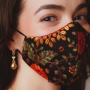 Op de afbeelding: Een vrouw draagt ​​een zwart mondkapje met een bloemenpatroon in tinten oranje, geel, groen en rood. Het masker heeft een zwart elastiek en is gemaakt van stof. De vrouw draagt ​​gouden oorbellen.