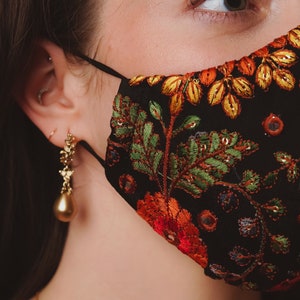Op de afbeelding: Een close-up van een persoon die een zwart gezichtsmasker draagt met een bloemenpatroon. Het masker heeft rode, oranje, gele, groene en blauwe borduurwerk. De persoon draagt gouden oorbellen.