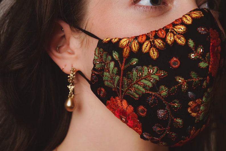 Op de afbeelding: Een close-up van een persoon die een zwart gezichtsmasker draagt met een bloemenpatroon. Het masker heeft rode, oranje, gele, groene en blauwe borduurwerk. De persoon draagt gouden oorbellen.