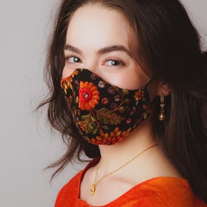 Op de afbeelding: Een vrouw met lang donker haar draagt ​​een zwart gezichtsmasker met een bloemenpatroon. Het masker heeft rode, oranje, gele, groene en blauwe bloemen erop. Ze draagt ​​ook een gouden ketting met een hanger en gouden oorbellen.