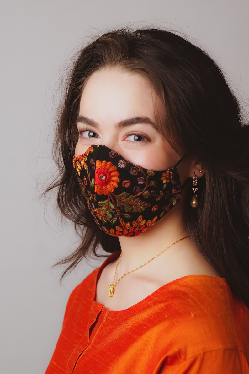 Op de afbeelding: Een vrouw met lang donker haar draagt ​​een zwart gezichtsmasker met een bloemenpatroon. Het masker heeft rode, oranje, gele, groene en blauwe bloemen erop. Ze draagt ​​ook een gouden ketting met een hanger en gouden oorbellen.