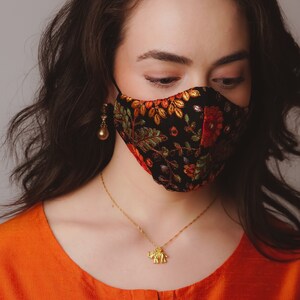 Op de afbeelding: Een vrouw draagt ​​een zwart mondkapje met kleurrijke bloemenborduurwerk. Ze draagt ​​een gouden ketting met een olifantenhanger en een paar gouden oorbellen.