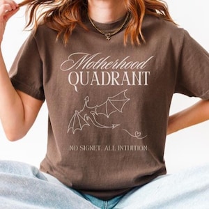 A model wears a comfort colors shirt in espresso brown. This is a shirt inspired by the Fourth Wing series. It says Motherhood Quadrant, No signet All intuition. It would be a perfect bookish gift for moms who love to read