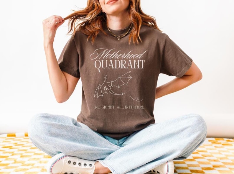 A model wears a comfort colors shirt in espresso brown. This is a shirt inspired by the Fourth Wing series. It says Motherhood Quadrant, No signet All intuition. It would be a perfect bookish gift for moms who love to read