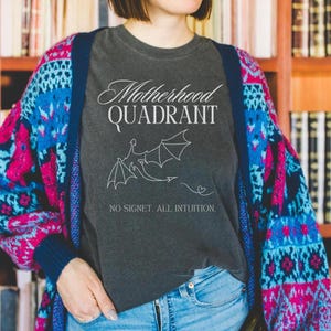 A model wears a comfort colors shirt in pepper. The Fourth Wing shirt is perfect for mothers who love to read. It says Motherhood Quadrant across the front and has a dragon and a plume of air that makes a heart