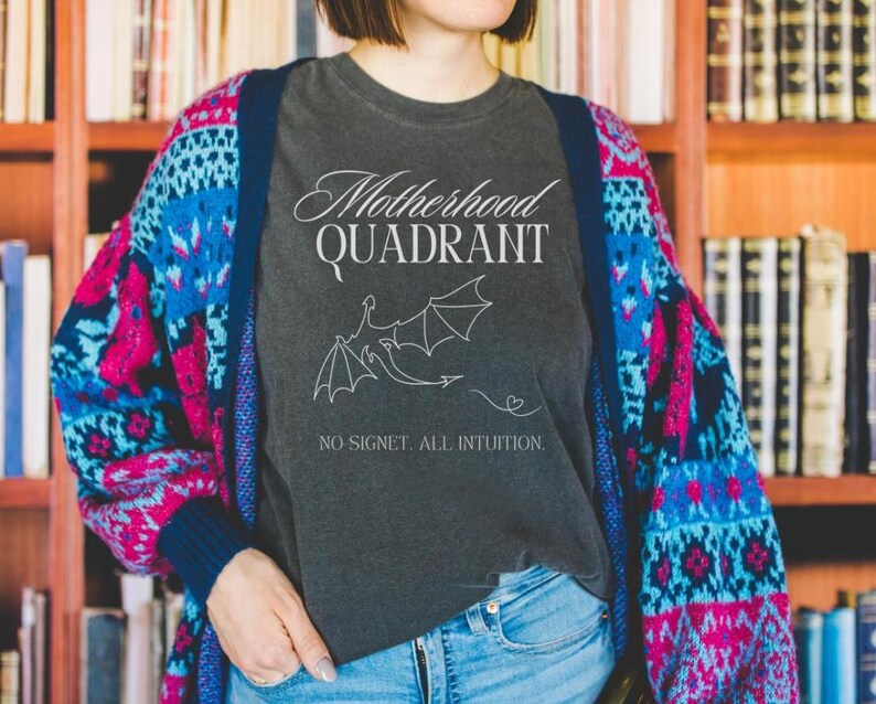 A model wears a comfort colors shirt in pepper. The Fourth Wing shirt is perfect for mothers who love to read. It says Motherhood Quadrant across the front and has a dragon and a plume of air that makes a heart