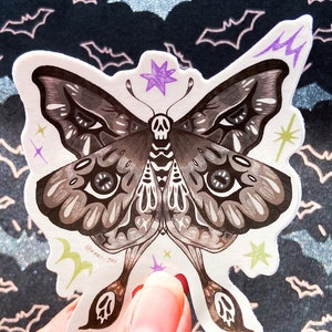 May include: A black and white moth sticker with a skull and crossbones design. The moth has large, detailed eyes and is surrounded by small stars and lightning bolts. The sticker is on a white background with a black and white bat pattern.