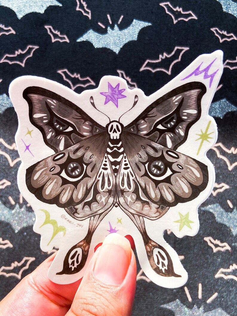 May include: A black and white moth sticker with a skull and crossbones design. The moth has large, detailed eyes and is surrounded by small stars and lightning bolts. The sticker is on a white background with a black and white bat pattern.
