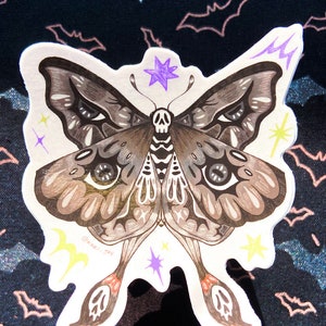 May include: A sticker of a detailed black and brown moth with a skull on its body. The moth has large eyes and is surrounded by yellow and purple stars. The sticker is on a black background with a pattern of bats.