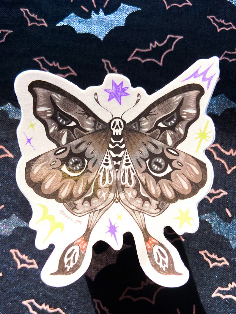 May include: A sticker of a detailed black and brown moth with a skull on its body. The moth has large eyes and is surrounded by yellow and purple stars. The sticker is on a black background with a pattern of bats.