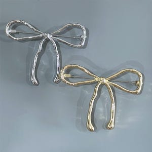 May include: Two bow-shaped brooches. One is silver-colored, the other is gold-colored. Both have a pin on the back for attachment. The bows have a textured, melted appearance, with elongated, curved tails.