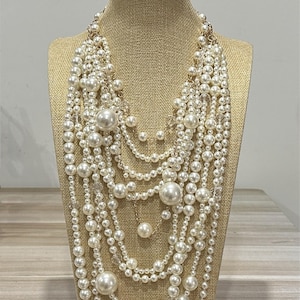 Könnte beinhalten: Mehrreihige Perlenkette auf einer Büste. Die Halskette besteht aus weißen Perlen in verschiedenen Größen, mit goldenen Kettenakzenten, die das Design ergänzen.