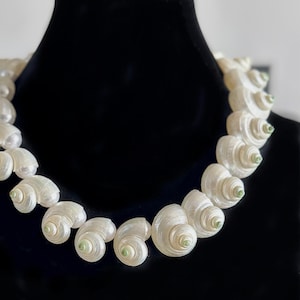 Op de afbeelding: Een witte schelpenketting met groene accenten. De ketting is gemaakt van natuurlijke schelpen en is aan elkaar geregen op een wit koord.