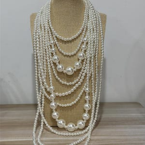 Puede incluir: Un collar de perlas de múltiples hilos exhibido en un maniquí. El collar presenta perlas blancas de varios tamaños, creando un efecto en cascada. Las perlas varían desde pequeñas perlas de semilla hasta cuentas más grandes, ofreciendo un aspecto clásico y elegante.