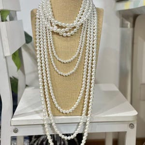 May include: A collection of white pearl necklaces of varying lengths displayed on a mannequin. The necklaces range from choker length to long strands, showcasing the different sizes and styles of pearls. The pearls have a luminous sheen.