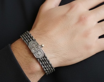 May include: A silver bracelet with a woven chain design. The bracelet has a decorative clasp with geometric patterns. The bracelet is worn on a wrist against a dark background. The bracelet is a piece of jewellery.