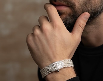 May include: A silver bracelet featuring a multi-strand woven chain design and a rectangular metal clasp. The clasp is decorated with intricate geometric patterns and a small central stud, providing a secure closure for this metallic wrist accessory.