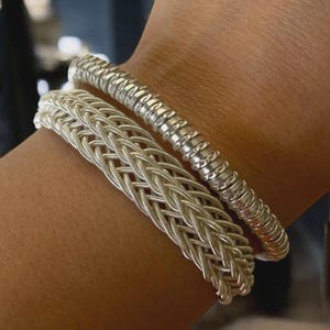 May include: Two silver bracelets are shown on a wrist. One bracelet features a woven pattern, and the other has a coiled design. The bracelets are made of a shiny, metallic material, reflecting light. These bracelets are a simple, elegant accessory.