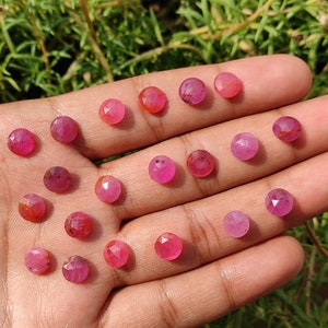 May include: A collection of 18 pink ruby gemstones, faceted and round, displayed on a hand. The gemstones are various shades of pink, from light to dark.