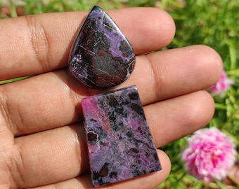 Op de afbeelding: Twee paarse en zwarte edelsteen cabochons, een traanvormig en een rechthoekig, worden op een hand getoond.