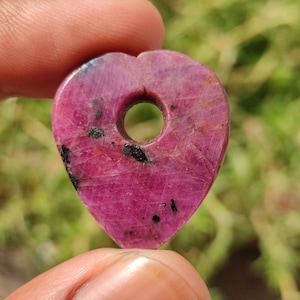 May include: A heart-shaped pink ruby pendant with a hole drilled through the top. The ruby has black inclusions throughout.
