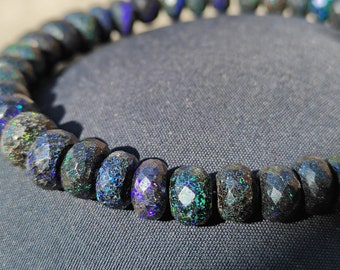 May include: A beaded bracelet featuring faceted, dark-colored beads with iridescent flashes of blue, green, and purple. The beads are strung together, forming a circular shape. The bracelet is displayed on a dark gray surface.