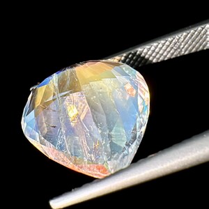 May include: A clear, faceted gemstone with rainbow iridescence. The gemstone is held by a pair of tweezers.