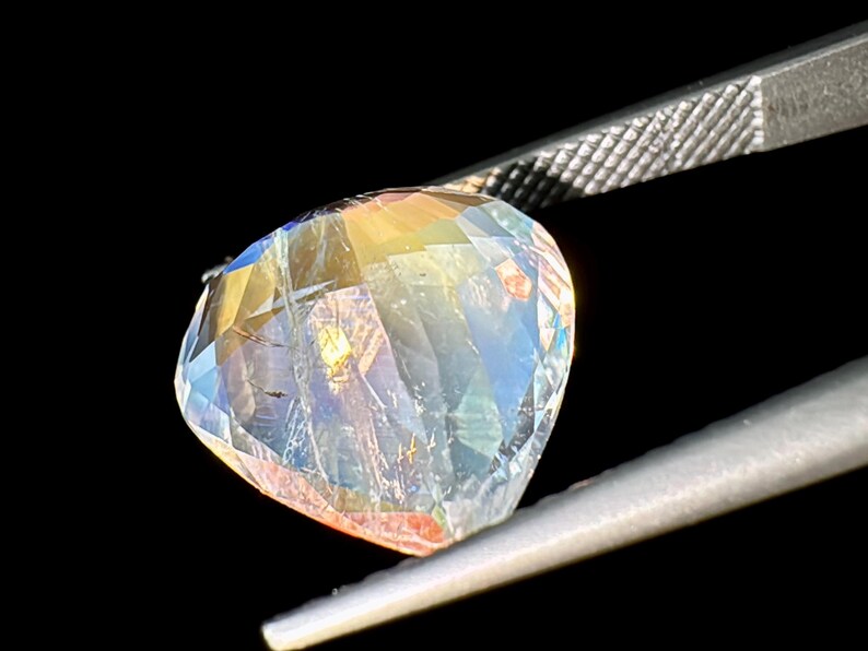 May include: A clear, faceted gemstone with rainbow iridescence. The gemstone is held by a pair of tweezers.