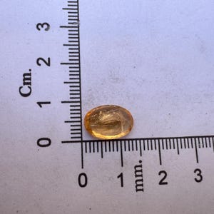 May include: A small, oval-shaped, translucent, amber-colored gemstone with a slight shimmer. The gemstone is approximately 1 cm long and is sitting on a white surface with a ruler for scale.