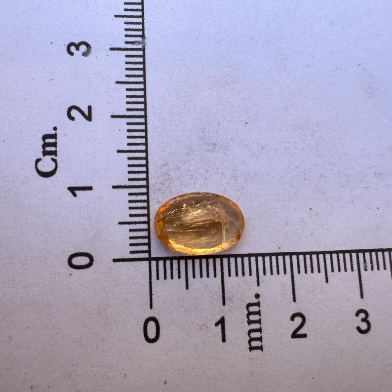 May include: A small, oval-shaped, translucent, amber-colored gemstone with a slight shimmer. The gemstone is approximately 1 cm long and is sitting on a white surface with a ruler for scale.