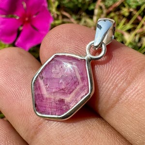 K&ouml;nnte beinhalten: Ein Anh&auml;nger mit einem polierten, unregelm&auml;&szlig;ig sechseckigen rosa Edelstein mit sichtbaren inneren B&auml;ndern und kristallinen Mustern, eingefasst in eine glatte, silberfarbene Metallfassung mit einer kleinen runden &Ouml;se oben.