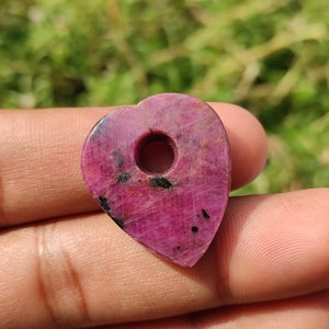 May include: A heart-shaped pendant made of pink ruby with a hole in the center. The pendant has a dark pink color with black inclusions.