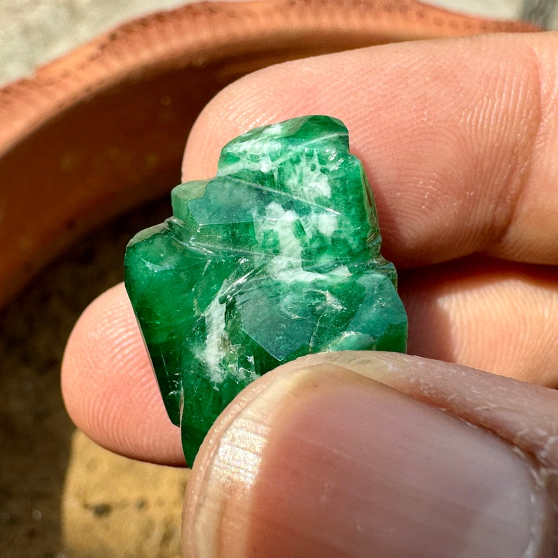 May include: A carved green emerald gemstone with white veins. The gemstone is held in a hand.