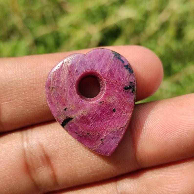 May include: A heart-shaped pendant made of pink ruby with black inclusions. The pendant has a hole drilled through the center for stringing.