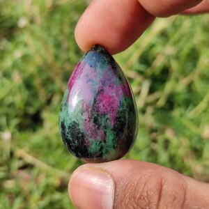 May include: A teardrop-shaped green and pink gemstone cabochon. The stone has a smooth, polished surface and is set in a silver bezel.