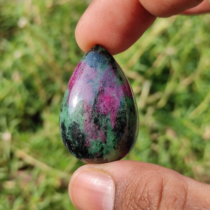 May include: A teardrop-shaped green and pink gemstone cabochon. The stone has a smooth, polished surface and is set in a silver bezel.