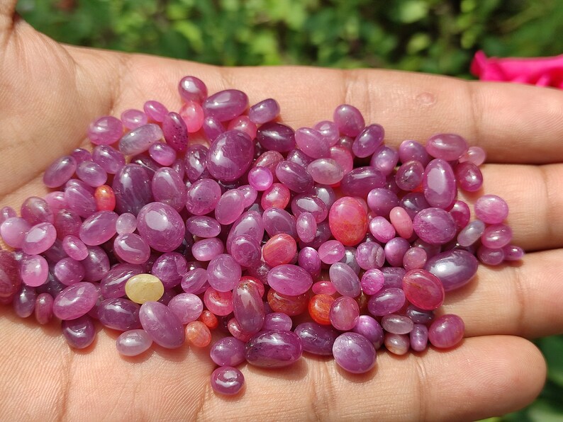 May include: A collection of polished, oval-shaped gemstones in varying shades of pink and red. The gemstones are smooth and glossy, with a range of sizes. The gems are held in a hand, with a blurred green background.