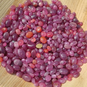 May include: A collection of polished, oval-shaped gemstones in shades of purple, pink, and red. The gemstones vary in size and are scattered on a light brown, textured surface. The gems have a smooth, glossy appearance.